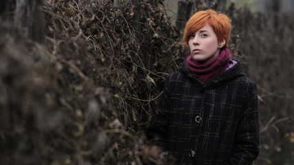 sweet red-haired girl in a black coat and purple knitted scarf is standing by the fence overgrown with grapevine or ivy, girl or woman on a background of autumn or spring nature, overcast