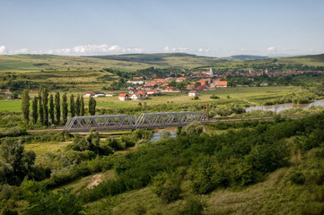 A steel train bridge over river in Romania with village in background and vegetation in foreground
