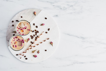 Iced coffee with rose and cardamom in a tall glass surrounded by coffe beans, dried roses and straws on white serving plate over white marble table. Top view with copy space for text.