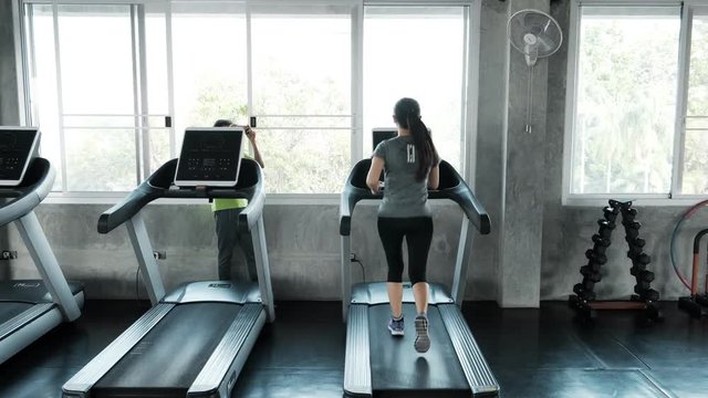 Senior woman using a running machine in gym, running slowly with her husband taking photo. Authentic Senior life style concept.