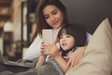 Love of young mother and daughter. Teaching reading a phone on the bed at home. they look at the phone on hand of girl which feel good and happy.