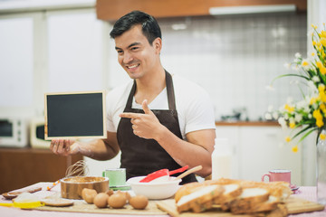 A man sitting which show a small black board. On the table have material for prepare dough powder. Chef have a aprons. Live at home in the kitchen. He smile.
