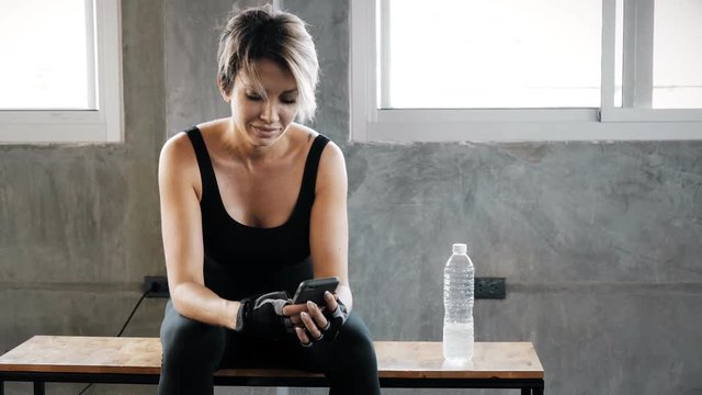 Authentic young caucasian woman using her mobile phone in gym to record and track her fitness performance, sitting on bench. Fitness mobile technology concept.