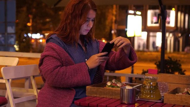 Young pretty red haired woman taking photo on her food at street cafe with beautiful night lights next to the road. 3840x2160, 4k