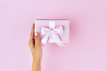Woman hands holding present box with pink bow on pastel pink background. Flat lay style.