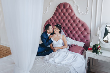 Obraz na płótnie Canvas Lovers and smiling newlyweds tenderly embrace on a bed in a modern studio. A chic portrait of a stylish groom with glasses and a beautiful bride in a white dress with a diadem. Wedding photography.