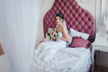 Obraz na płótnie Canvas A beautiful bride in a white dress with a bouquet is sitting on a large bed in a modern studio and smiling. Chic portrait of a cute brunette. Wedding photography.