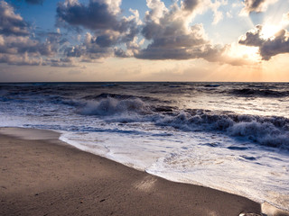 Beach at sunset with choppy sea and waves crashing to shore.