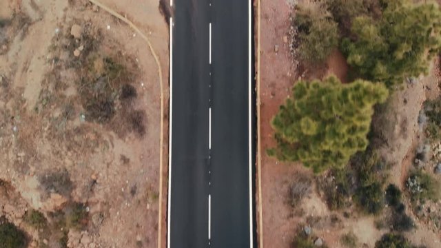 Aerial shot. Looking down. Cars go new ideally flat asphalt road. Fresh white road markings. Against the background of green trees and red stone desert landscape. Spain, Canary Islands, Tenerife