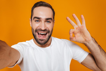 Portrait of unshaved man 30s in t-shirt smiling while taking selfie photo, isolated over yellow...