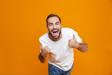 Image of joyous guy 30s in t-shirt rejoicing and showing thumbs up while standing, isolated over yellow background