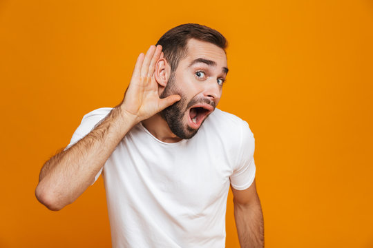 Image of positive man 30s trying to hear something while keeping hand at his ear, isolated over yellow background