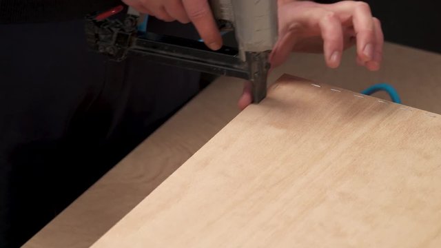 A view from top of a man's hand holding pneumatic staple gun and stapling a plywood board to wooden drawer