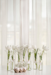 White tulips in bottles with Easter decoration in the form of eggs on a white table.
