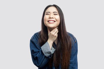 Throat pain. Portrait of sick brunette asian young woman in casual blue denim jacket with makeup standing and touching her painful throat. indoor studio shot, isolated on light grey background.