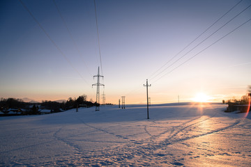 Electrical tower on a snowy field. Sun goes down, orange colors, evening scenery