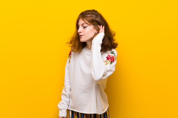 Young redhead woman over yellow wall listening to something by putting hand on the ear