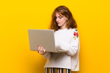 Young redhead woman over yellow wall with laptop