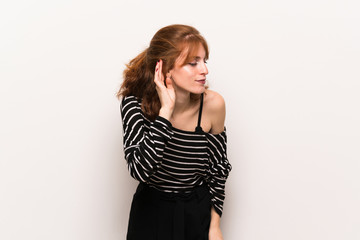 Young redhead woman over white wall listening to something by putting hand on the ear