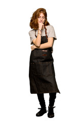 A full-length shot of a Young redhead woman with apron looking down with the hand on the chin over isolated white background