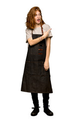 A full-length shot of a Young redhead woman with apron pointing to the side to present a product over isolated white background