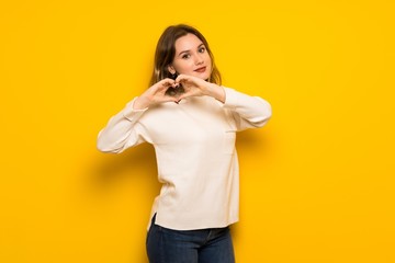 Teenager girl over yellow wall making heart symbol by hands