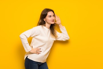 Teenager girl over yellow wall listening to something by putting hand on the ear