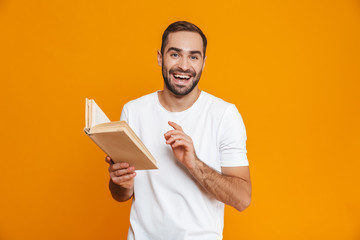 Obraz na płótnie Canvas Image of smiling man 30s in white t-shirt holding and reading book, isolated over yellow background