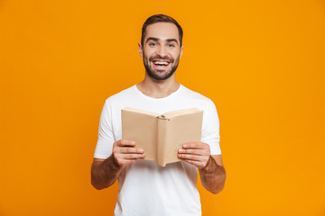 Image of optimistic man 30s in white t-shirt holding and reading book, isolated over yellow background