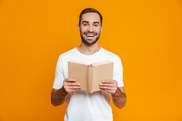 Image of optimistic man 30s in white t-shirt holding and reading book, isolated over yellow...
