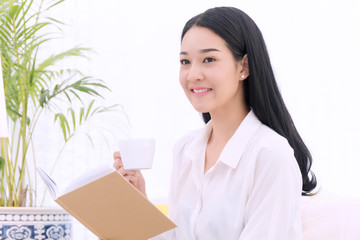 Cheerful smiling young woman reading a diary or book sitting on white sofa with holding a white cup of coffee in her hand at home. Beautiful girl black long hair looking at camera in living room.