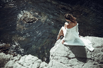 beautiful dark-haired young woman in white light dress with long legs sits at the edge of a rock above the sea, surrounded by sea and cliffs , back view