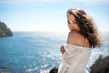 Fototapeta na wymiar Portrait of a beautiful young woman with dark fluttering hair dressed in white light dress standing at the edge of a rock and looking at camera, showing her shoulder, and blue sky and sea background