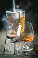 Glass and a bottle of brandy or cognac and smoking cigar on the wooden table.