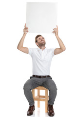 seated happy casual man holds blank board above his head