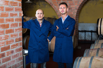 Fototapeta na wymiar two calm men in uniforms standing in cellar with wine woods