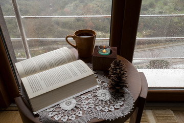 The cup of tea or coffee with a book and candle on the wooden table near the window on the rainy weather.