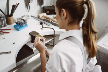 Focused on work. Side view of young female jeweler making a ring at her workbench. - obrazy, fototapety, plakaty