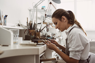 Focused on a process. Portrait of young female jeweler focused on creating a silver ring at her modern workbench. - obrazy, fototapety, plakaty