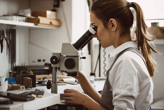 Utmost precision. Close up portrait of female jeweler looking at the new jewelery product through microscope in a workshop.