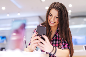 Happy brunette standing in tech store and trying out smart phone she want to buy.