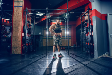 Young bearded Caucasian serious bodybuilder standing in the gym with hands on hips. Weight west on.