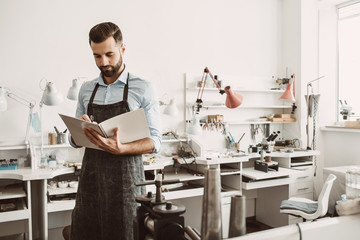 Report preparation. Portrait of bearded male jeweler in apron planning a day in notebook