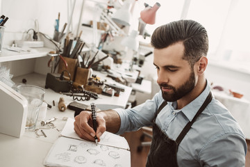 Creative work. Portrait of young bearded jeweler drawing a sketch of a new ring