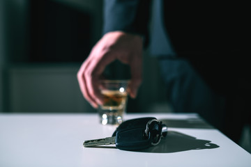 Close up of car keys on the desk. In background businessman taking shot of alcohol. Hotel room interior.