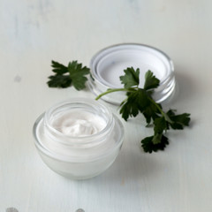 Obraz na płótnie Canvas Small jar with cream for skin care of eyelids and fresh green leaves of parsley on a light wooden background. Selective focus.