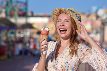 Young smiling woman in a wonderful dress and sunhat is eating an ice-cream in the Luna park. She is enjoying her holidays.