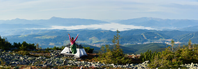 Panoramic view of camping on the top of mountain on bright summer morning. Back view of camper woman in pink sweater and sleeping bag standing in the entrance of tourist tent, holding hands lifting up
