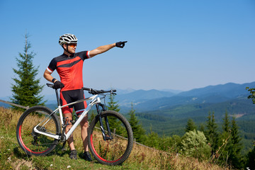 Fototapeta premium Young athletic professional biker standing with bicycle on top of hill, pointing into distance, enjoying view mountains, blue summer sky on background. Copy space. Active lifestyle and sport concept