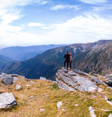 Fototapeta premium Back view of tourist couple, man and girl stand on rocky mountain top enjoying breathtaking autumn mountain panorama. Tourism, traveling and healthy lifestyle concept. motorcyclists, square photo